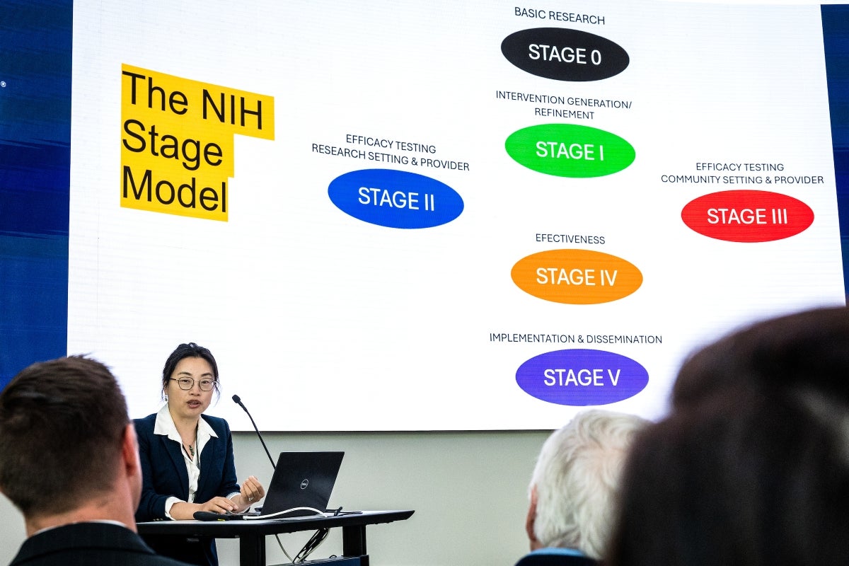 A woman speaks into a microphone in front of a screen displaying 'The NIH Stage Model.'