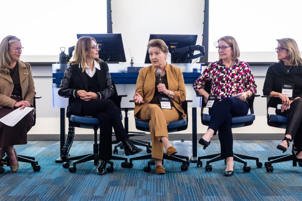 Five women sit in chairs in the front of a large room of people