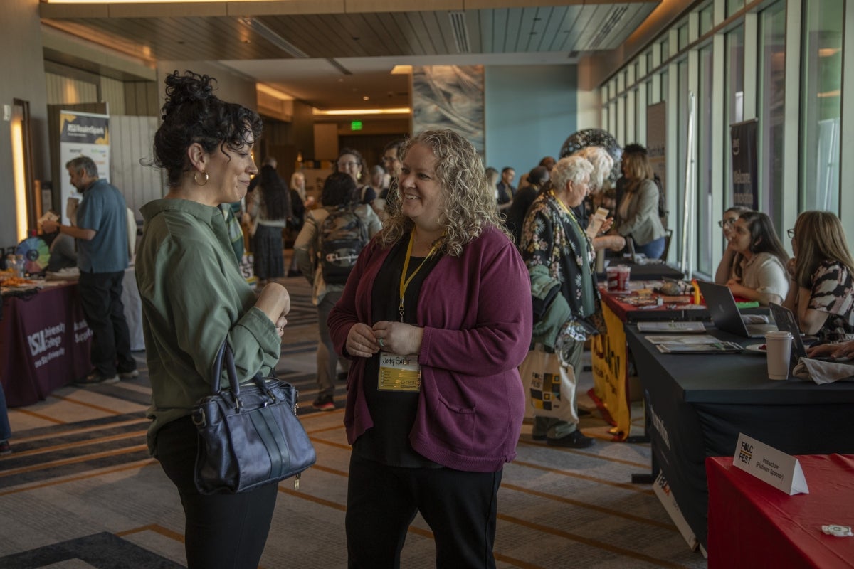 Two women speak in a hallway lined with various groups tabling.