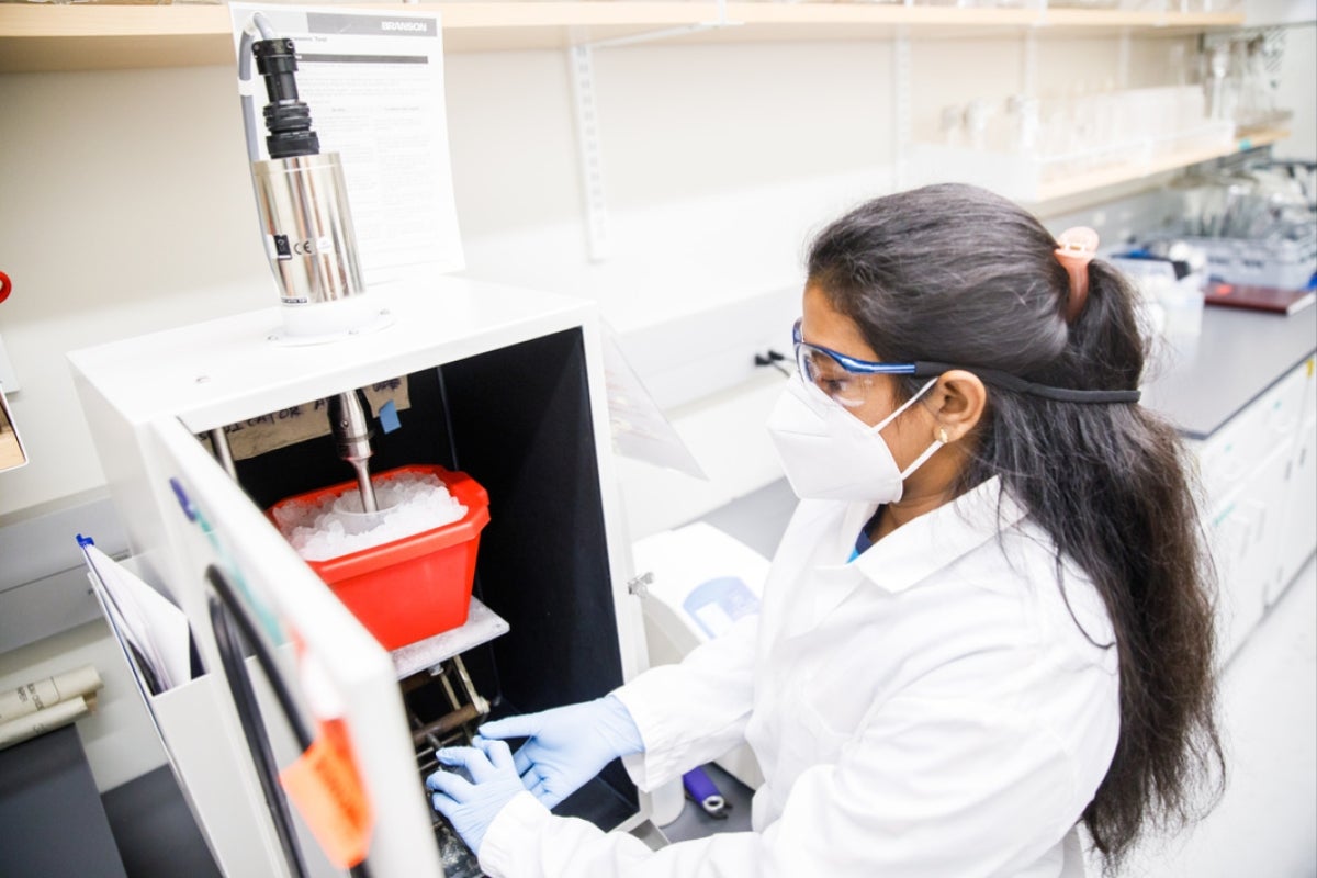 Woman wearing a white coat, face mask and goggles in a lab. She is working with a piece of machinery.