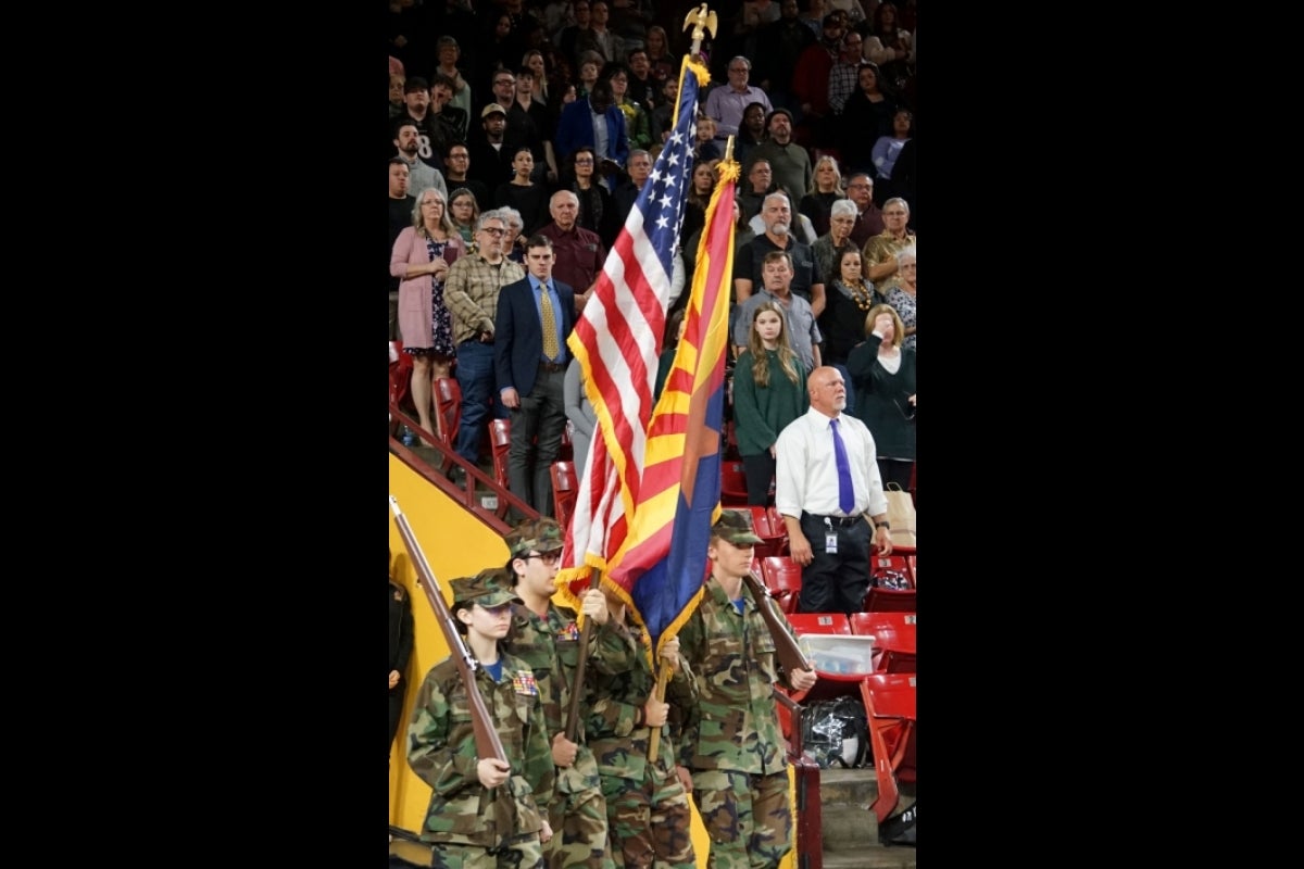 People dressed in Army gear holding US and Arizona state flags walk into a full stadium.