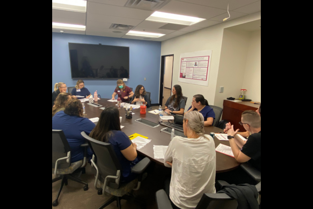 A group of students are seated at a conference table listening to directions. They all have a piece of paper in front of them with the scenarios they will be running through.