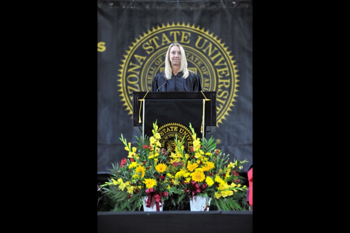 Kristine Kassel, ASU graduate '91 BS, wearing a black robe and speaking behind a lectern in front of the Arizona State University seal.