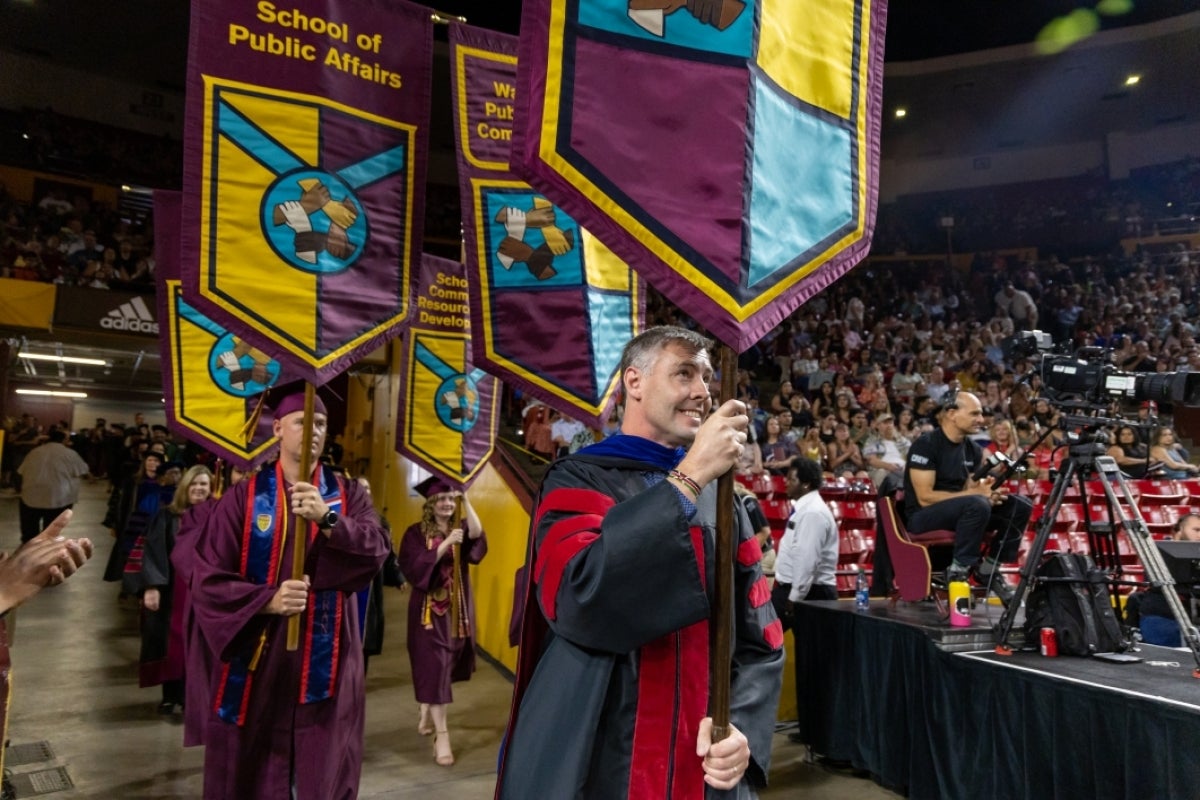 Associate Professor Eric Legg carries the Watts College gonfalon followed by others in graduation gowns.