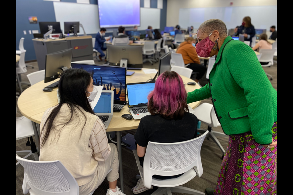 Students seated at a table with laptops as a professor leans in to instruct them.