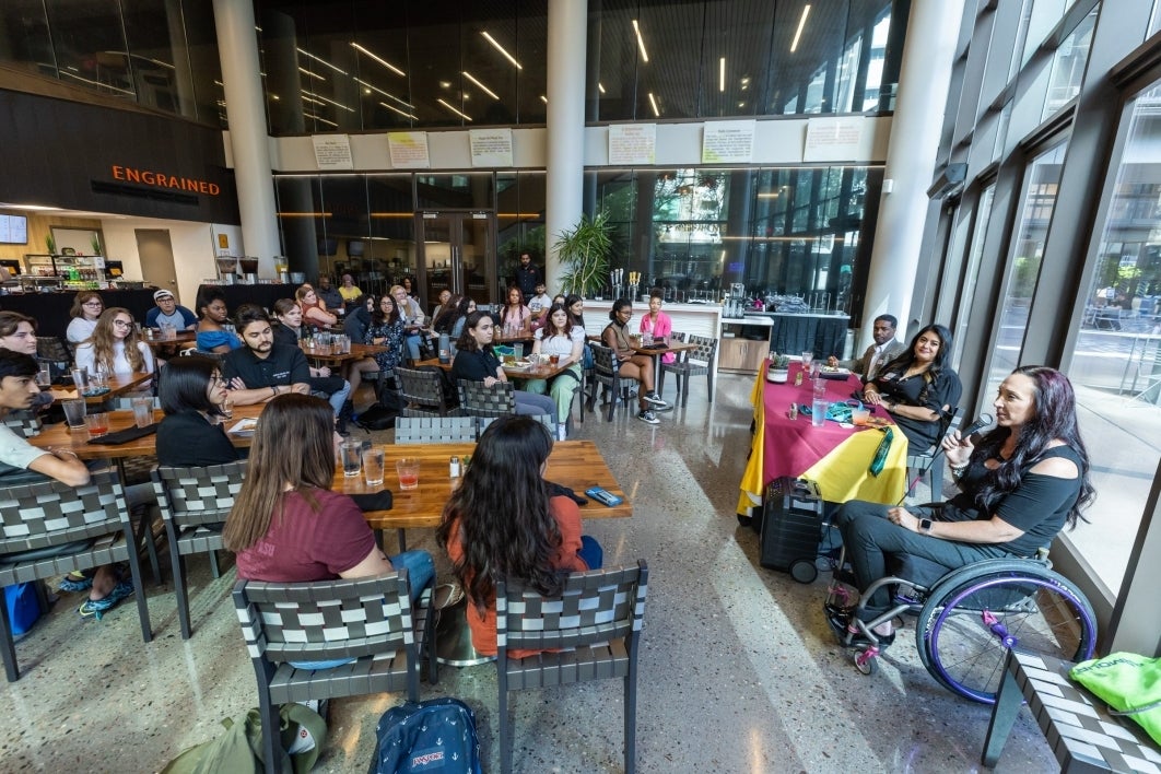 People seated at tables in a cafe listening to a woman seated in a wheelchair speaking into a microphone.