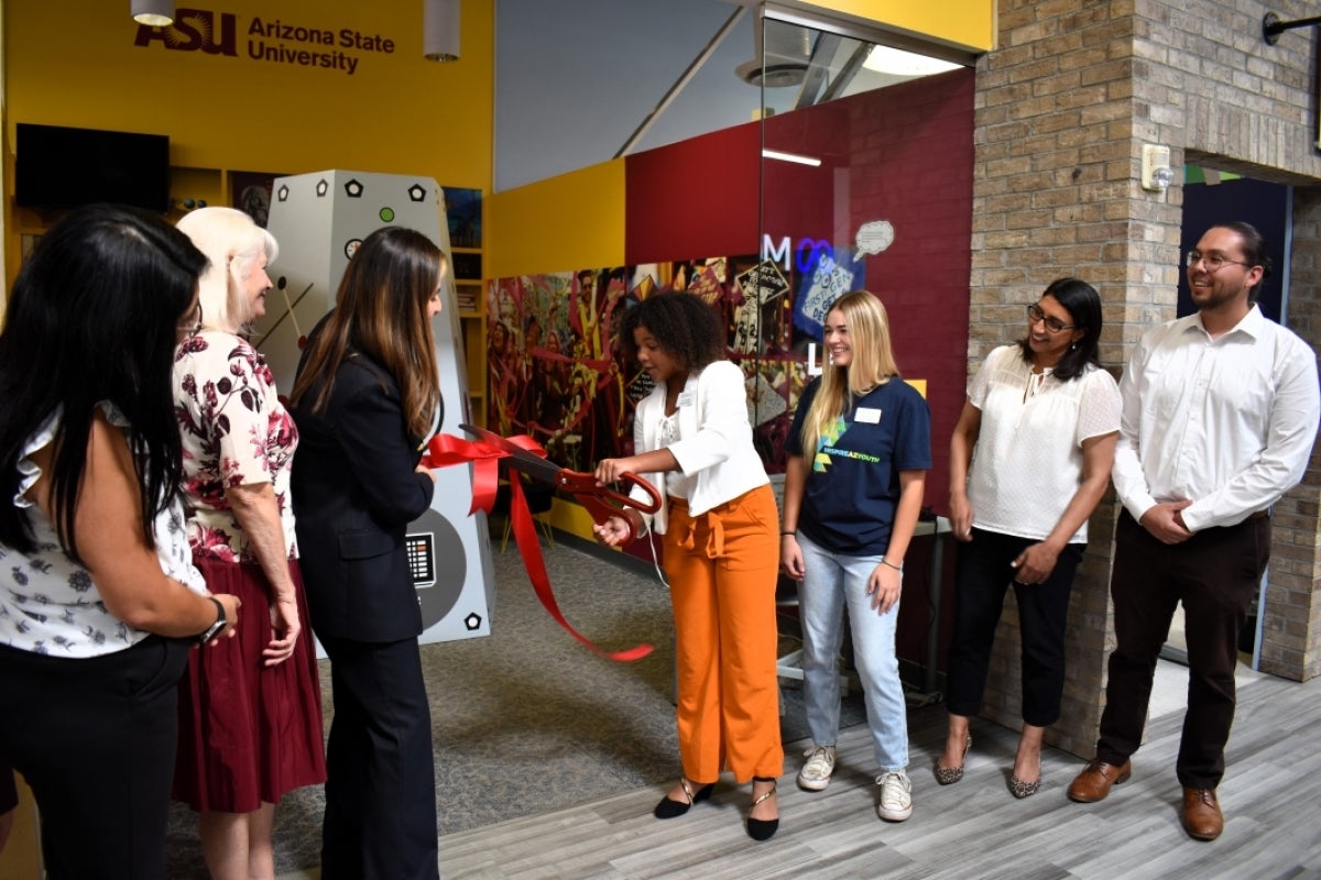 Young student cuts a large ceremonial ribbon as a crowd of people look on.