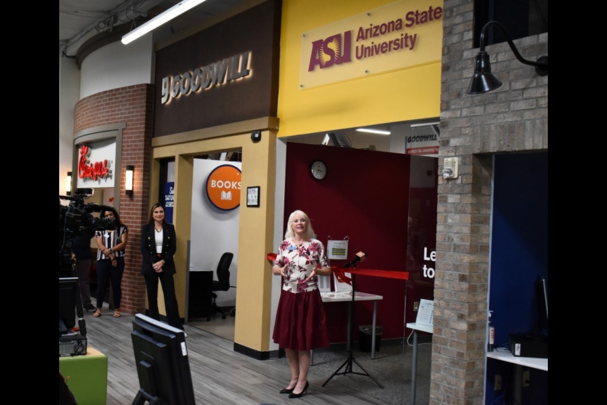 Woman speaking to a crowd in front of a storefront with a sign that reads 