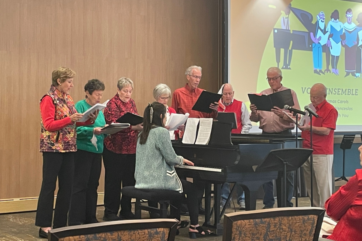 Student sitting at a piano as Mirabella residents surround her while looking at music sheets.