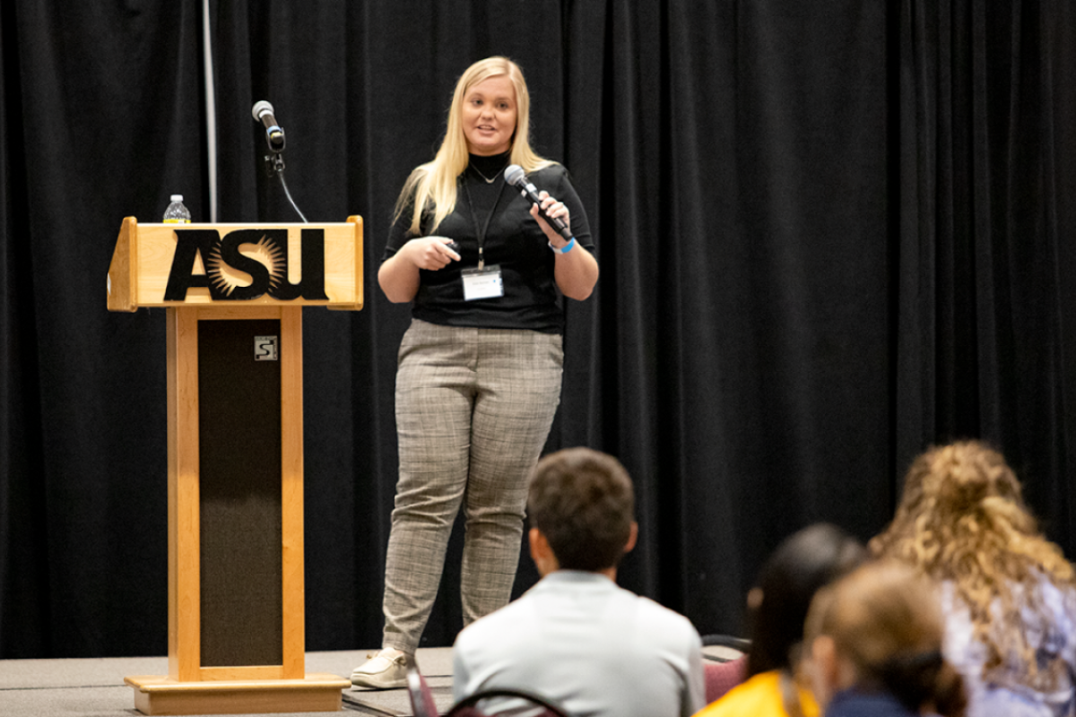 Woman standing on a stage speaking into a microphone.