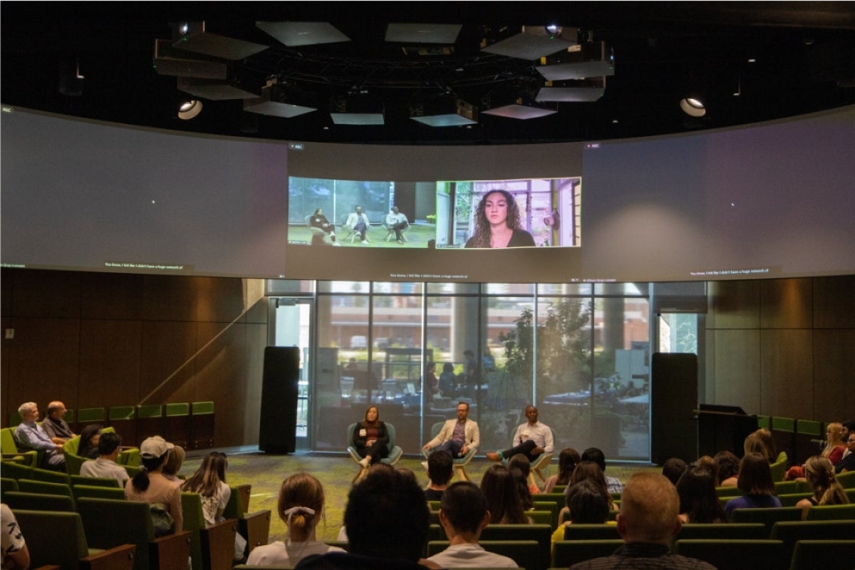 People seated onstage in a large auditorium with a large screen above them showing a woman speaking from a remote location.