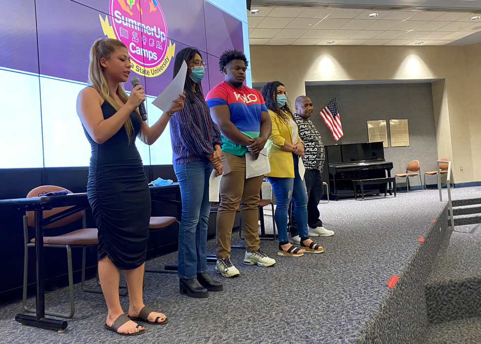 Students standing on a platform while one reads from a peice of paper and speaks into a microphone.