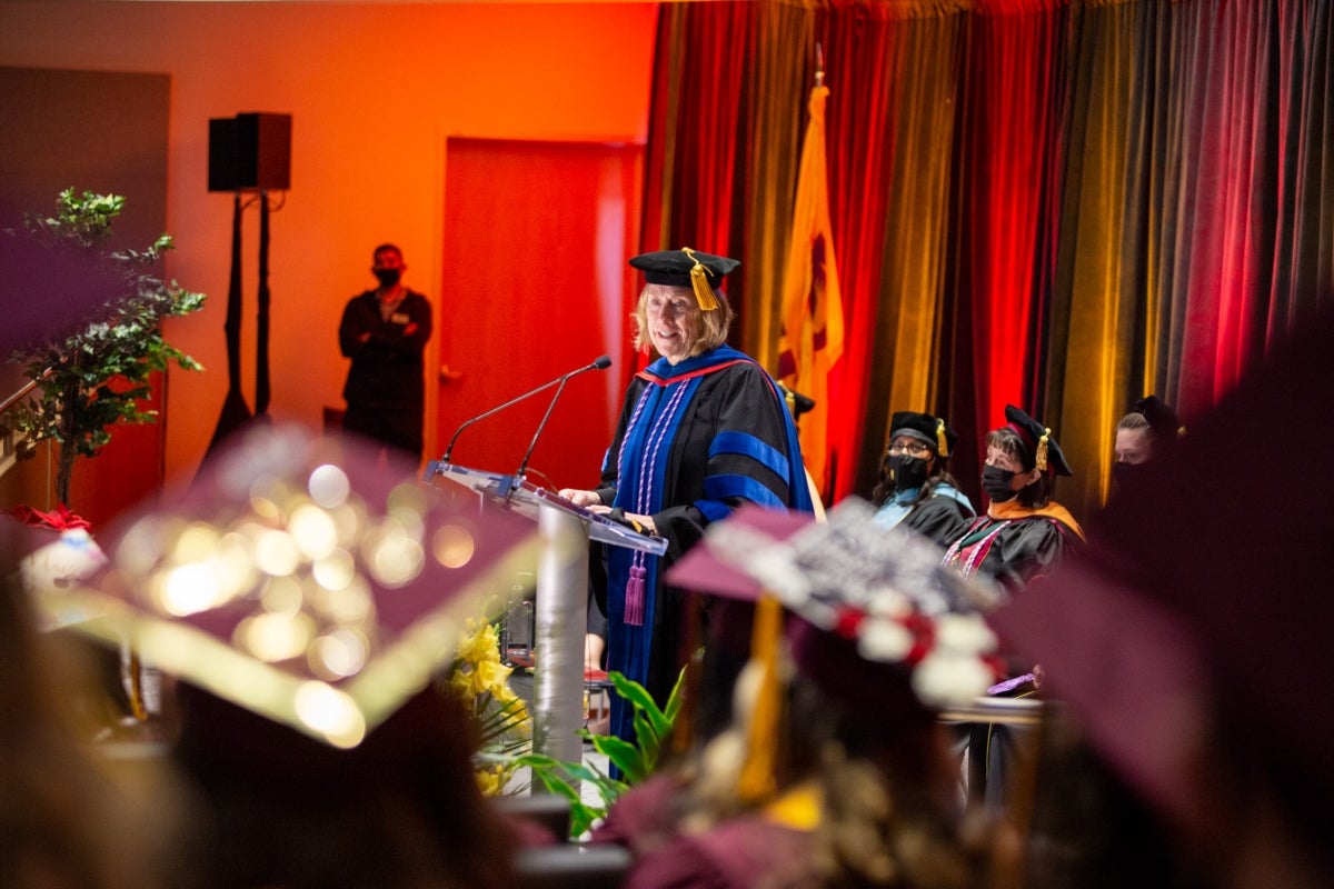 Dean Judith Karshmer addresses graduates. She's wearing a black gown with pops of blue and a black graduation cap with a gold tassel.