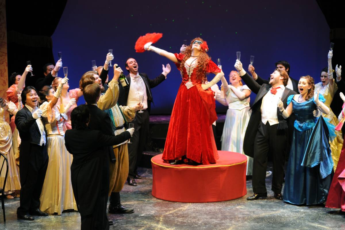 A woman in a red dress surrounded by other performers in opera attire.