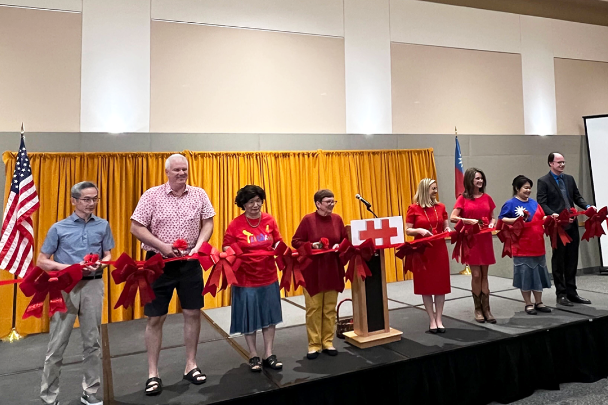 People standing on a stage in a row, holding a long red ribbon, with an American flag behind them.