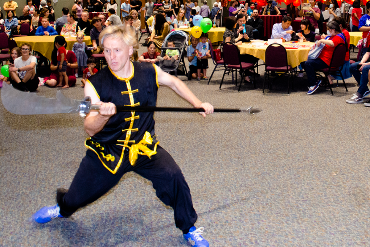 Man mid-movement, wearing a black outfit with gold trim, holding a large spear-like weapon as a crowd looks on.