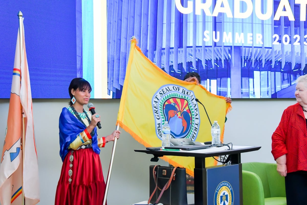 Cheyenne Gordon stands holding the ​​Hualapai Tribe flag in one hand a microphone in the other.