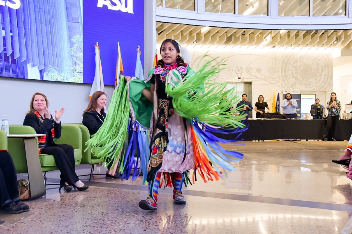 A woman in a colorful tribal outfit walks through a room with those around her clapping.