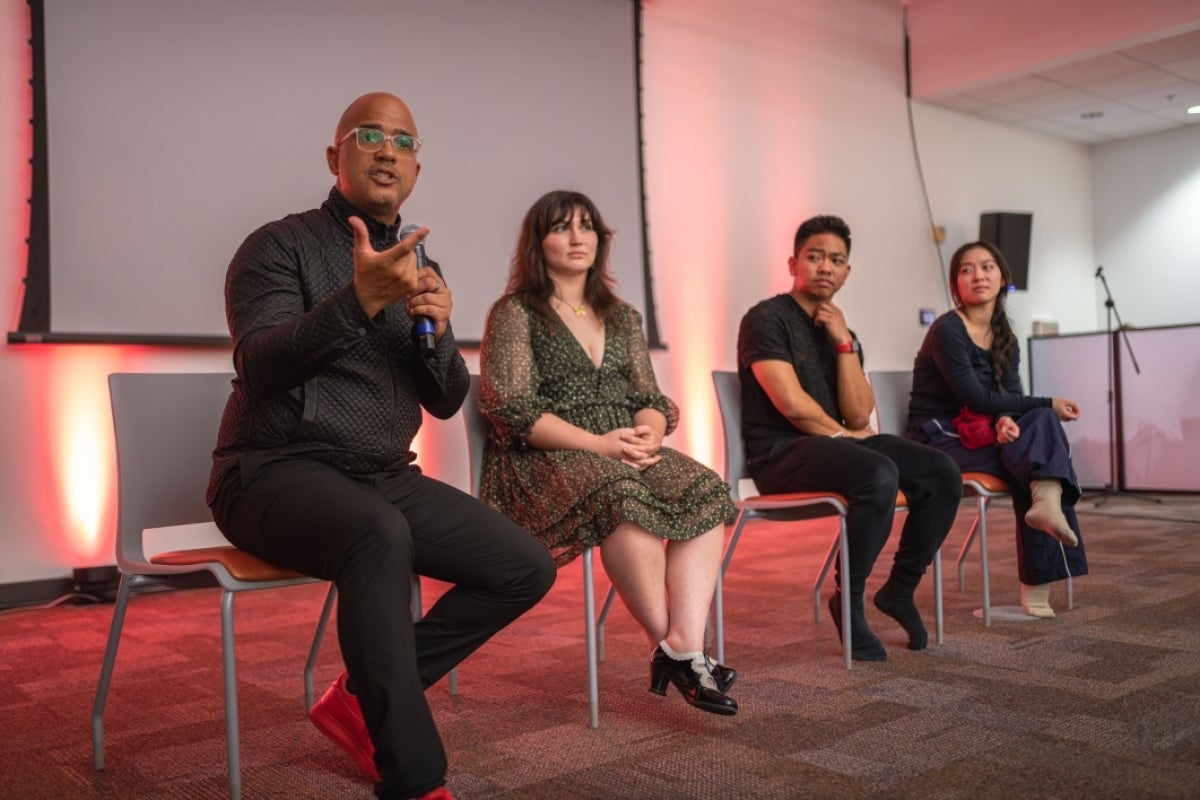 Four people seated on a stage listening to one as they speak into a microphone.