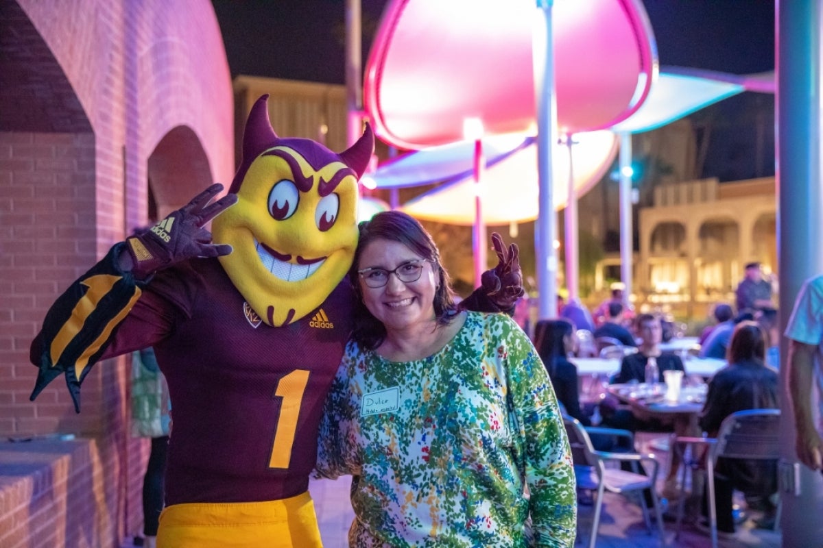 ASU Principal Lecturer Dulce Gonzáles-Estévez smiling and posing with Sparky the Sun Devil.