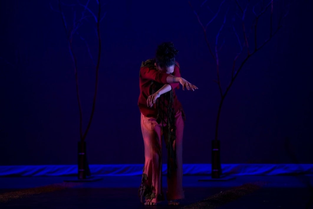 A performer wearing red is resting their head in their crossed arms against a dark, moody blue backdrop