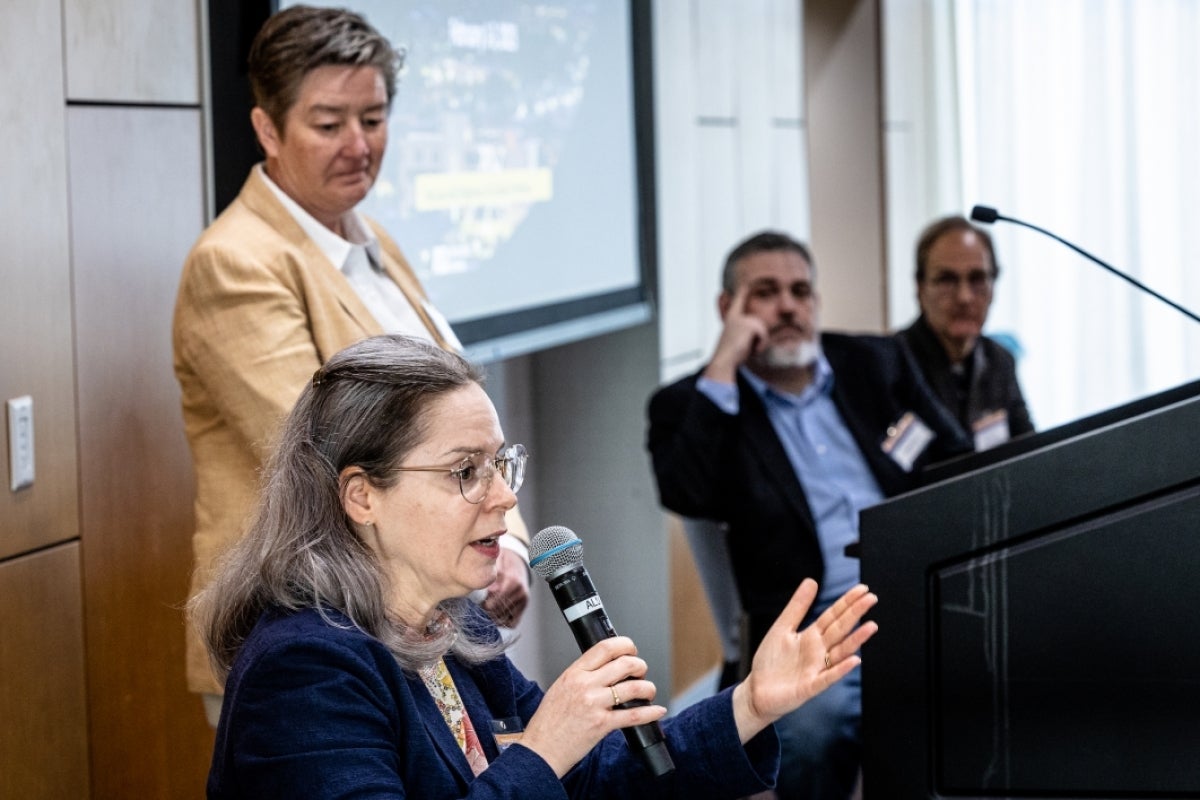 Woman speaking into a microphone while seated with other panel members behind a table.