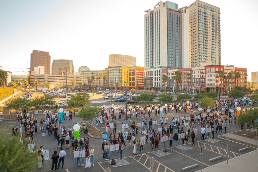 Hundreds of students stand by posters in the parking lot of a Downtown Phoenix building. In the background are skyscrapers