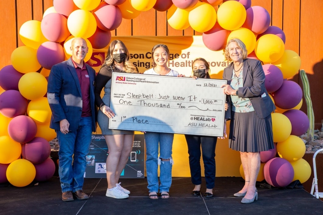 Three students hold a large check with on stage with a balloon backdrop behind them