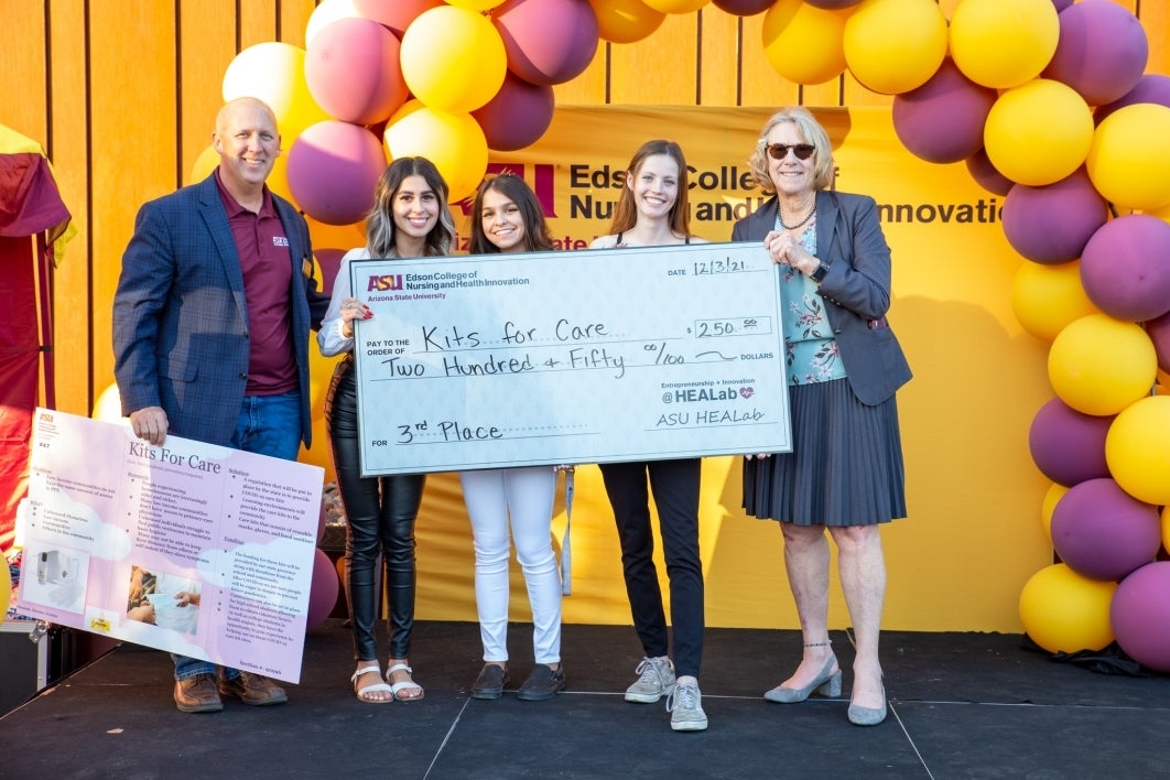 Three students hold a large check with on stage with a balloon backdrop behind them