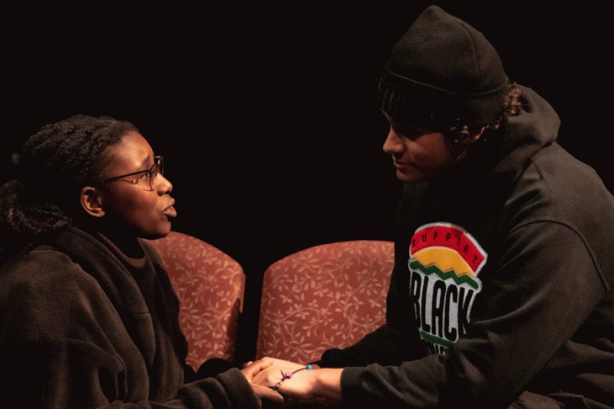 A man seated on a couch and a woman kneeling next to him hold hands on stage while acting in a play.