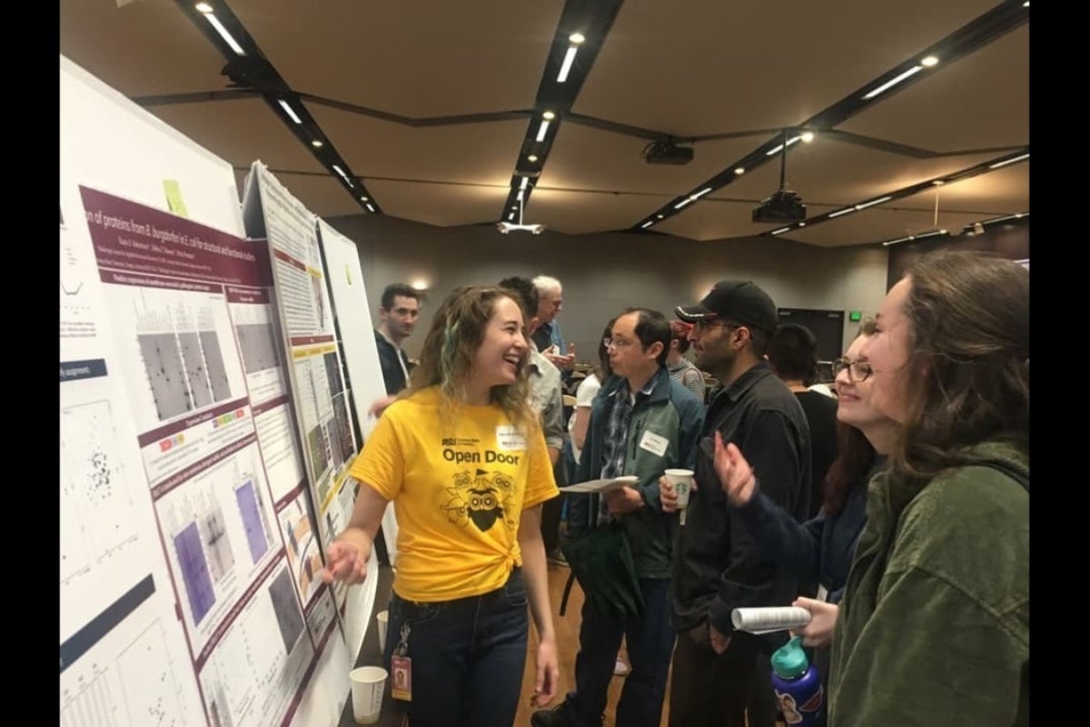 Woman smiling and pointing to a research poster as she speaks to onlookers.