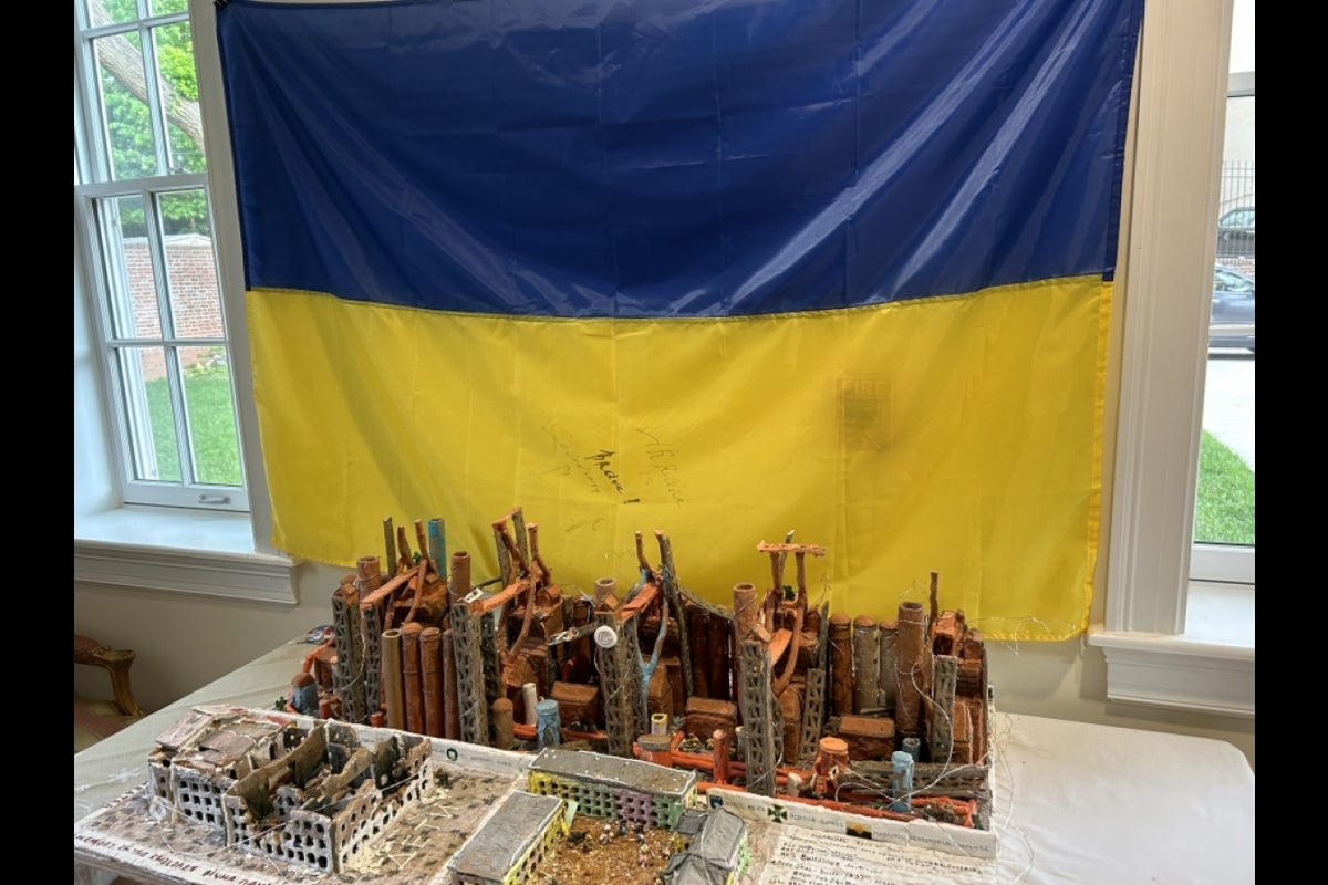 The Ukrainian flag hanging by a window with artwork of city ruins on a table in the forefront.