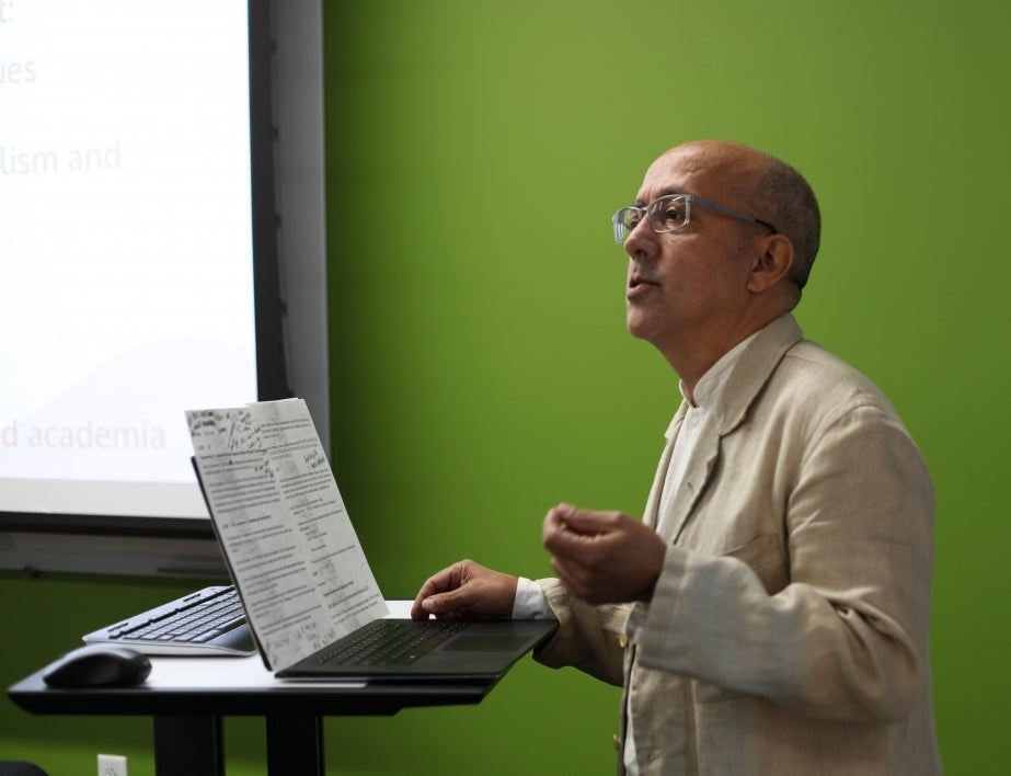 Jaime Fatas-Cabeza presents his research. He is wearing a tan suit coat over a white shirt. He is wearing glasses. Behind him is a neon green wall and a projector screen. He is holding his hands in the air as he talks.