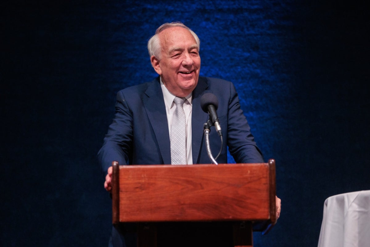 A man in a navy suit speaks at a wooden podium.