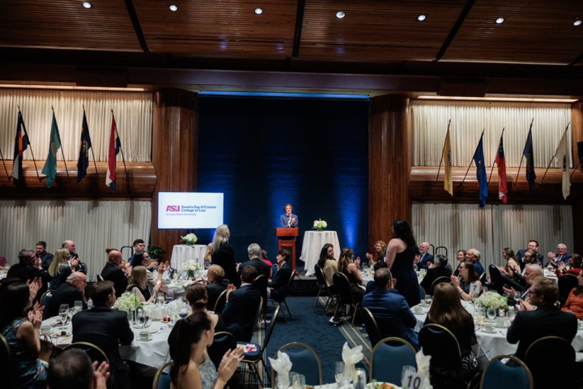 A crowd of guests fills the audience at white tables while a person speaks at a brown wooden podium.