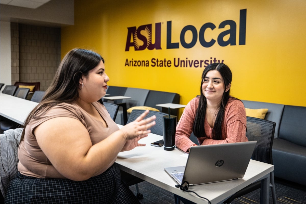 Two students seated in a classroom talking to each other. In the background, a sign on a wall says 