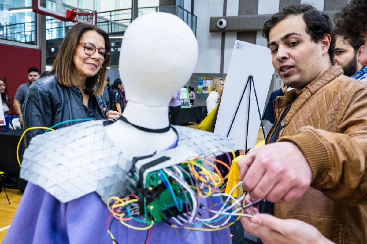 Woman looks at a mannequin wearing a superhero cape with an electrical device on it.