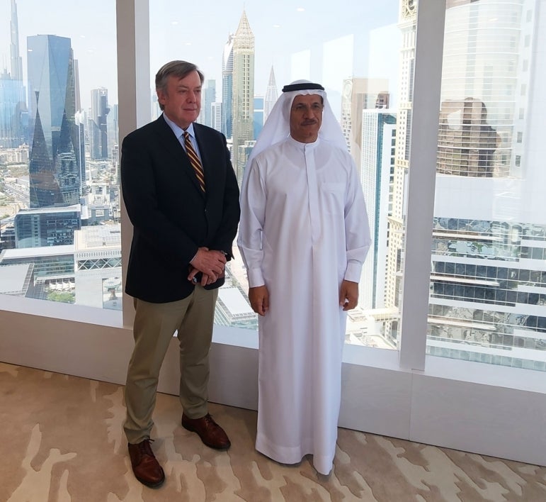 Two men pose for a photo in front of a city skyline, one man in Western suit and the other in traditional Arab dishdasha