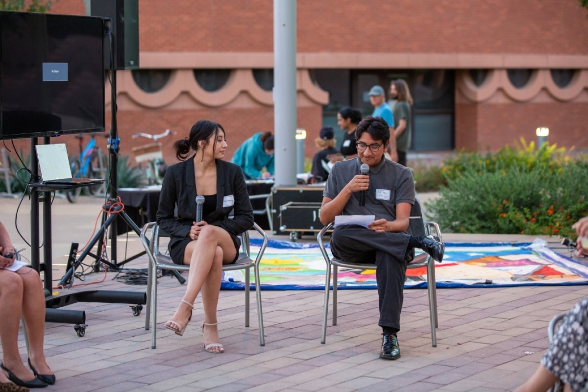 Two people seated next to each other holding microphones at an outdoor event.