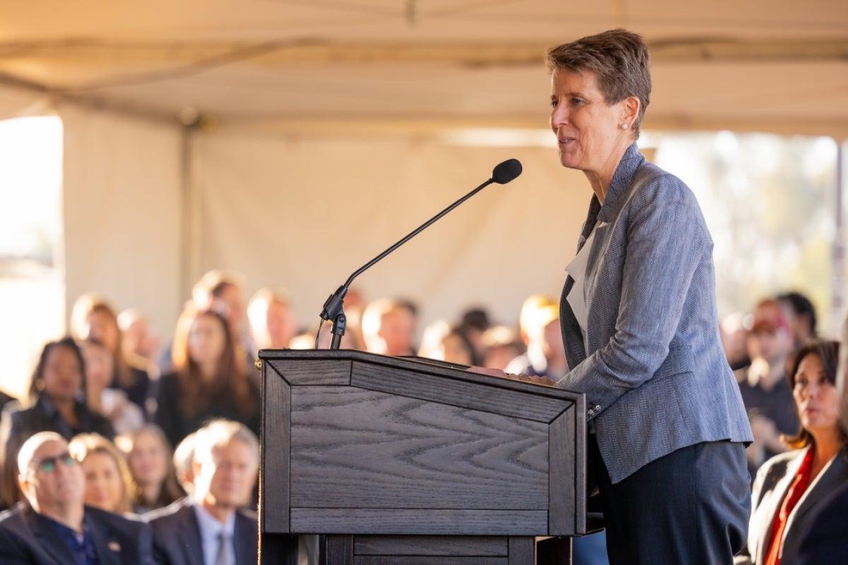 Woman speaking into microphone behind a lectern to a crowd.