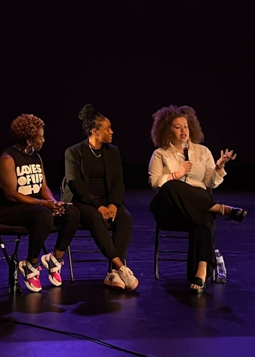 Three people sit on chairs next to one another onstage as one speaks into a microphone.