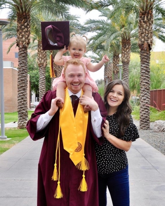 ASU College of Health Solutions graduate Jacob Nicoll smiles and poses with his family, his daughter perched on his shoulders and holding his graduation cap, on Palm Walk at ASU's Tempe campus