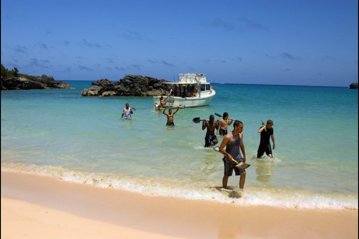 Students holding shovels and equipment walk from a boat docked near a shore onto a beach.