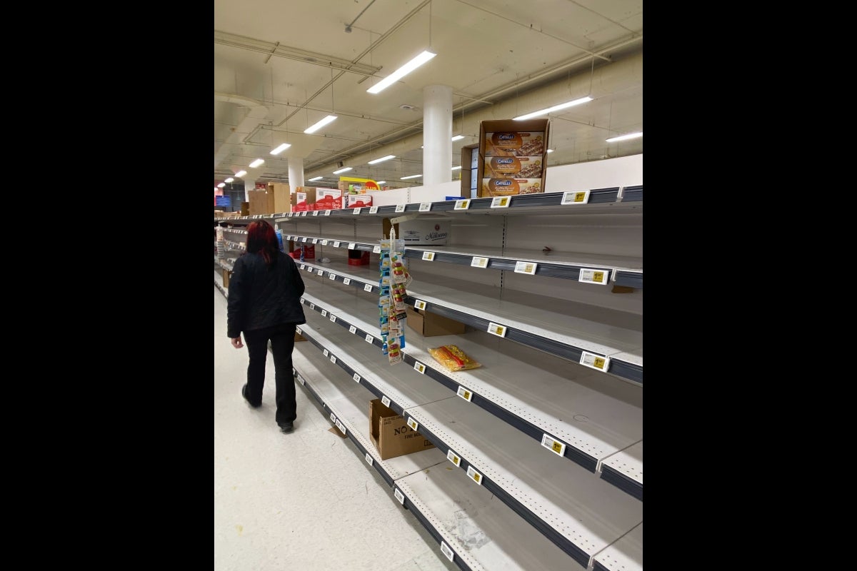 Person seen from the back, walking down an aisle in a grocery store with empty shelves.