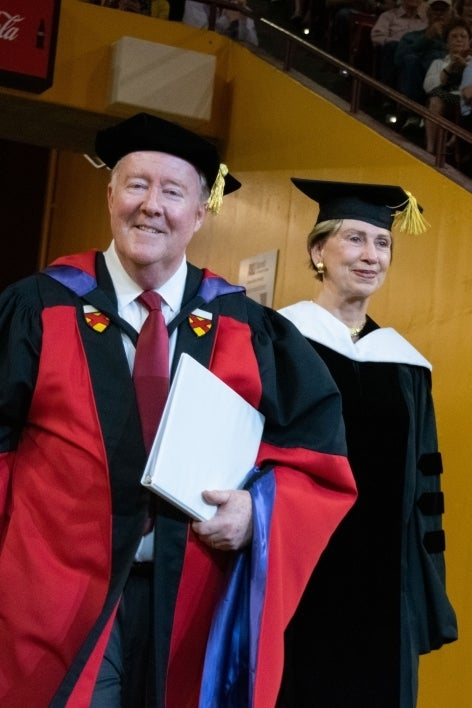 A man and a woman in graduation regalia walk into an arena.