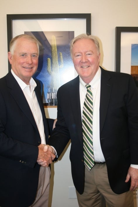 Two men shake hands in an office while smiling at the camera