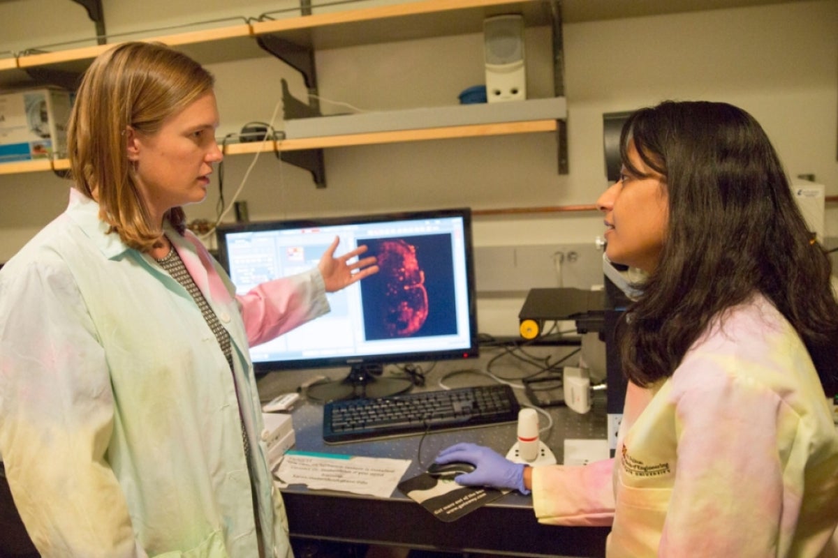 Sarah Stabenfeldt and Vimala Bharadwaj discussing research in a lab with a computer screen showing a brain scan in the background