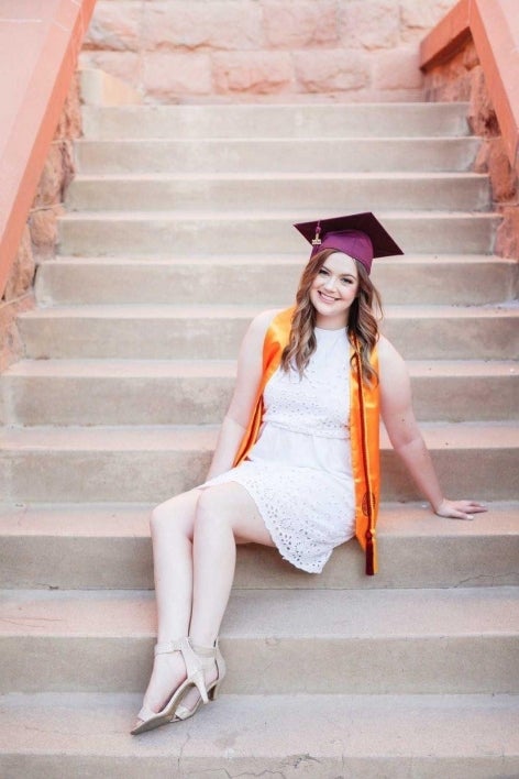 ASU's College of Health Solutions graduate Paige Snyder smiles in a white dress, wearing her graduation cap and sash while sitting on a staircase