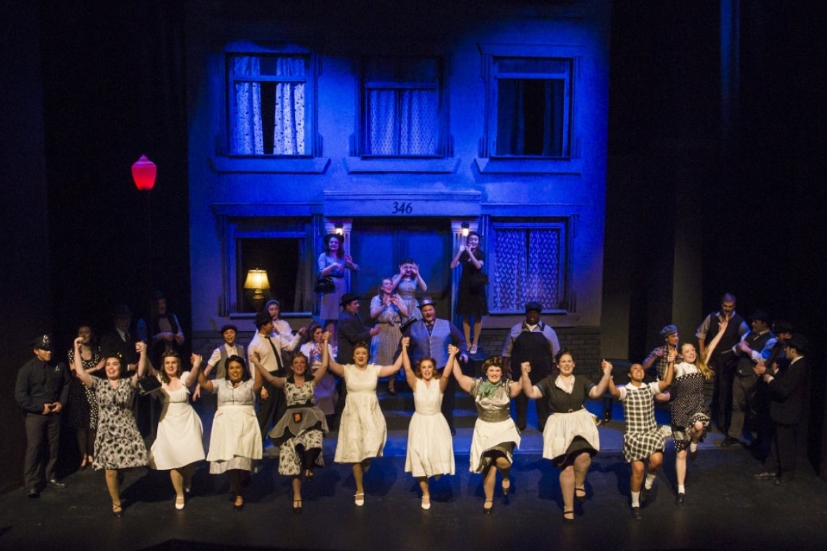 A group of performers in costume during a show with a staged house behind them.