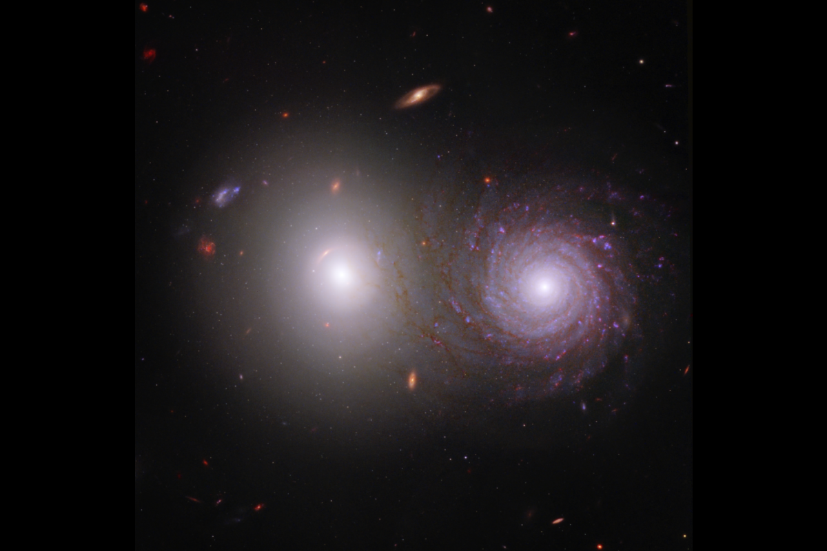 The majority of the image shows the black background of space. Two large, very bright galaxies dominate the center of the image. The elliptical galaxy at left is extremely bright at its circular core, with dimmer white light extending to its transparent c